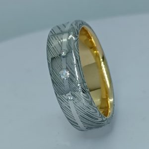 Une alliance en acier de damas, 6 mm de large, intérieur en or jaune 750/1000, fil en or blanc ondulant /7501000 serti de 3 diamants